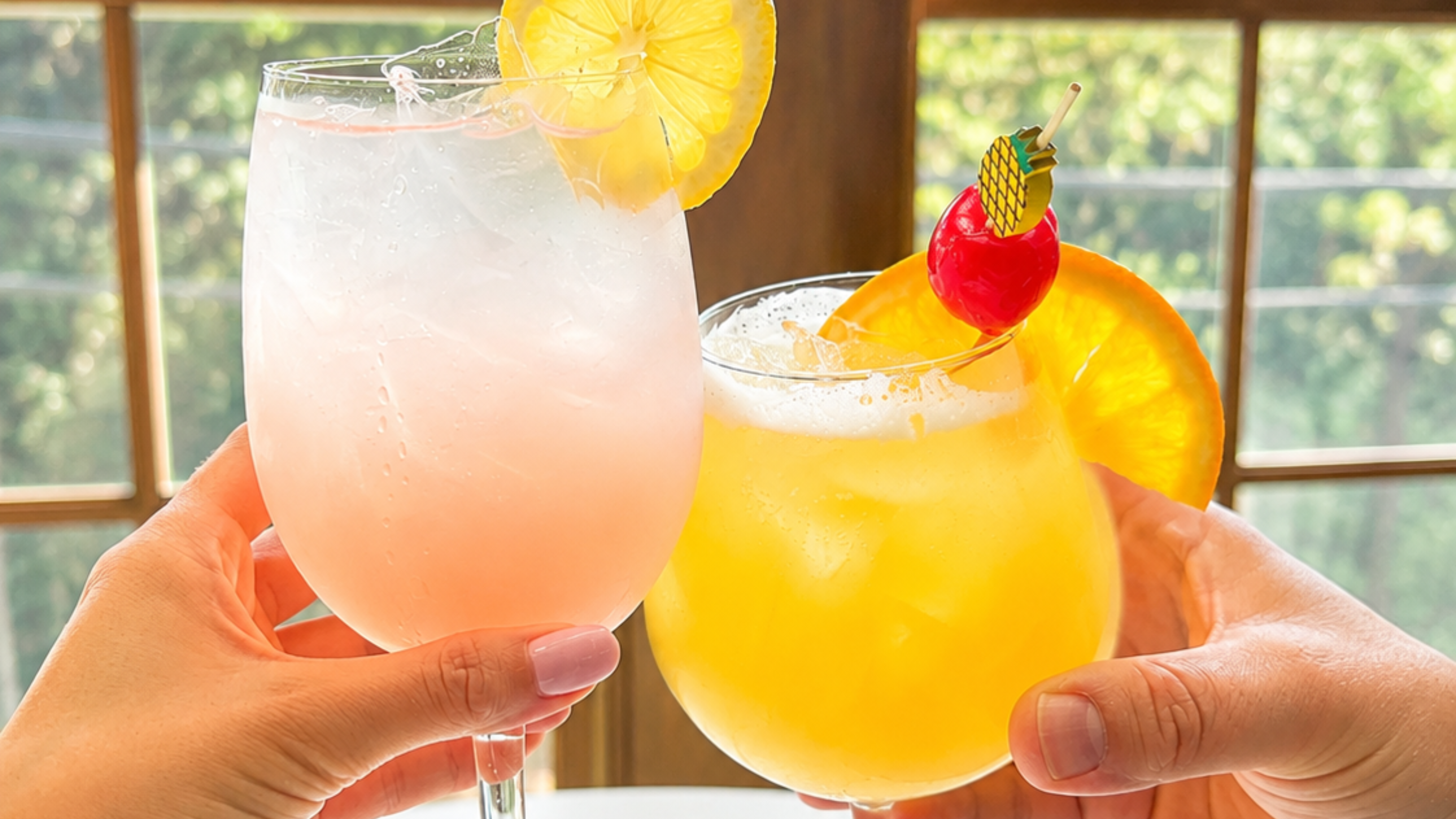 Two hands clink two citrus cocktails—pink lemonade and bright yellow drink—topped with fruit slices and playful straws.