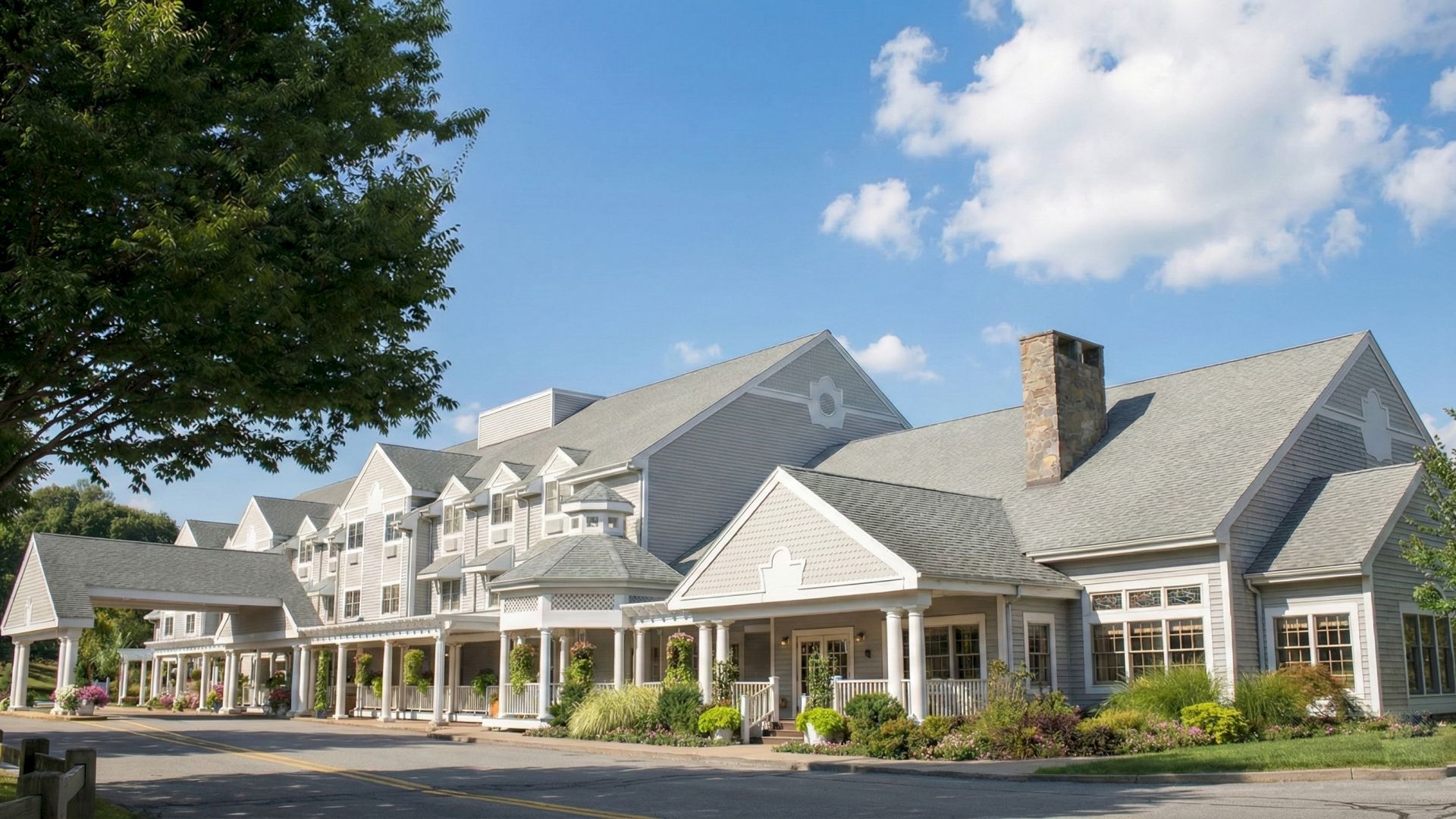 A large, elegant hotel or resort building with gray roofs, white siding, a covered entrance, and manicured landscaping under a bright blue sky with clouds.