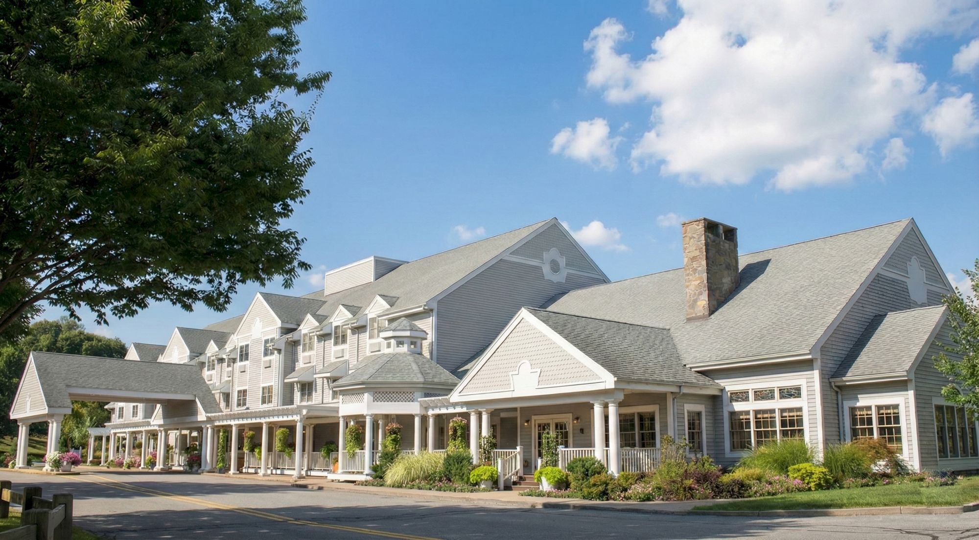 A large, elegant hotel or resort building with gray roofs, white siding, a covered entrance, and manicured landscaping under a bright blue sky with clouds.