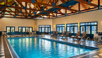 An indoor swimming pool with tiled deck, lounge chairs, and blue steel rafters under a vaulted wooden ceiling.