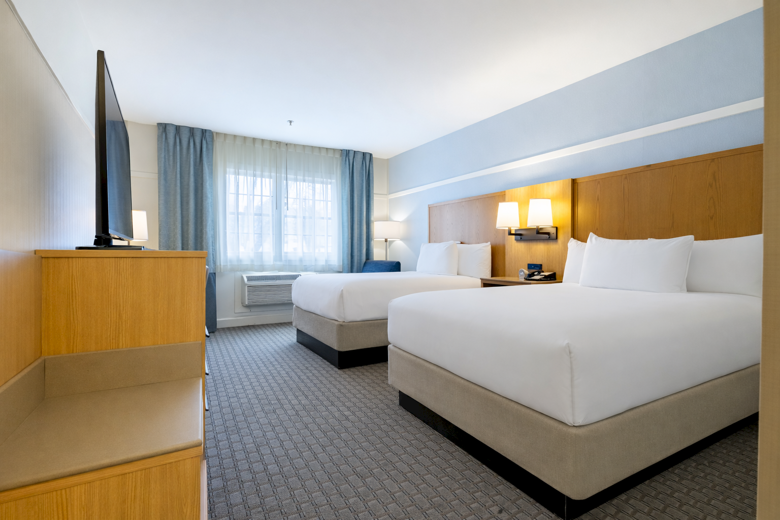 A modern hotel room with two neatly made twin beds, bedside lamps, and blue curtains by a window.