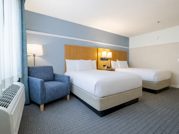 A hotel room with two queen beds, a blue accent wall, a blue armchair, a nightstand with lamps, and a window with sheer curtains, tidy and bright.