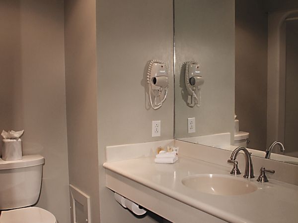 A small bathroom with a toilet and a long countertop sink area, mirror and wall-mounted hairdryers, gray tiles, and soft overhead lighting.