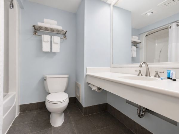 A modern bathroom with a toilet, a long white vanity sink, a large mirror, and a towel rack above the toilet; light blue walls and dark tiled floor.