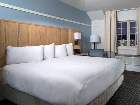 A hotel room with a large bed, white linens, wooden headboard, nightstands, a lamp, a chair, and a window with curtains.