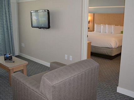 A hotel suite living area with a wall-mounted TV, gray sofa, coffee table, and a doorway leading to a bedroom with a king bed.