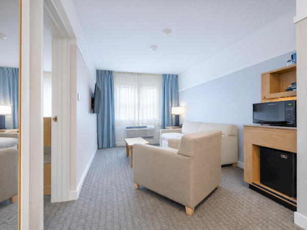A neat hotel living area with two beige armchairs, a small table, a desk with a microwave and mini-fridge, and a window with blue curtains.