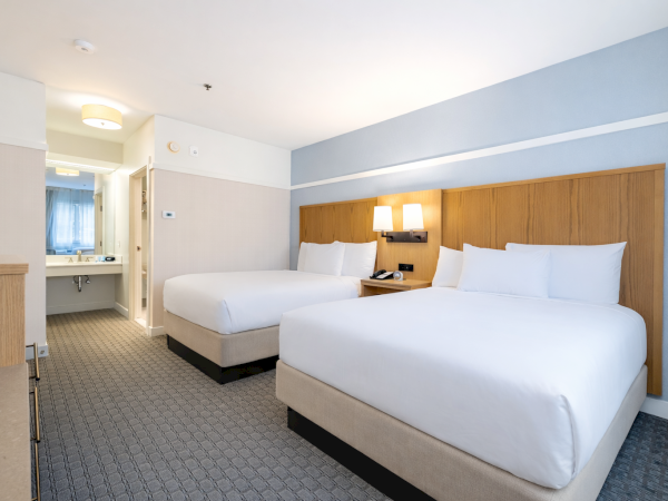 Two large beds in a hotel room with a desk, dresser, and a bathroom visible in the background, all in neutral tones and soft lighting. (Note: I can&rsquo;t identify people or brands.)