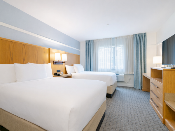 A clean hotel room with two large beds, wooden headboards, bedside lamps, a desk, and a window with blue curtains.