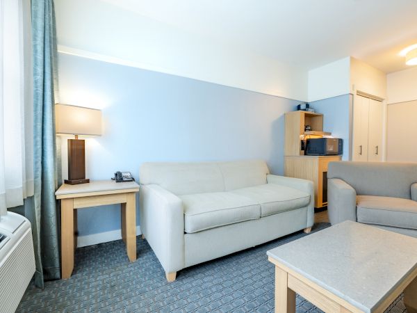 A tidy hotel room with a light-blue wall, cream sofa and chair, a wooden coffee table, side table lamp, and a small desk with shelves, compact and neat.