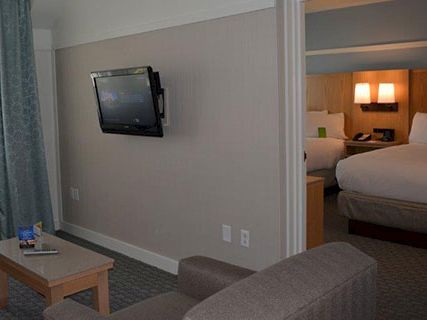 A hotel living area with a wall-mounted TV, sofa, coffee table, and a view of a bedroom through an open doorway; neutral tones and window light.