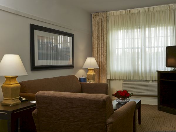 A cozy hotel living room with two brown armchairs, a sofa, side tables with lamps, a framed picture, a TV on a stand, and a window with sheer curtains.