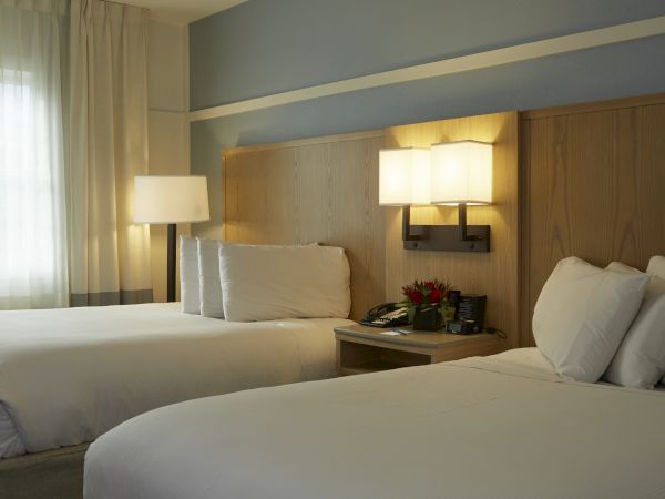 Two neatly made hotel beds with white linens, a nightstand, lamps, and soft natural light from a window, creating a calm, tidy bedroom ambiance.