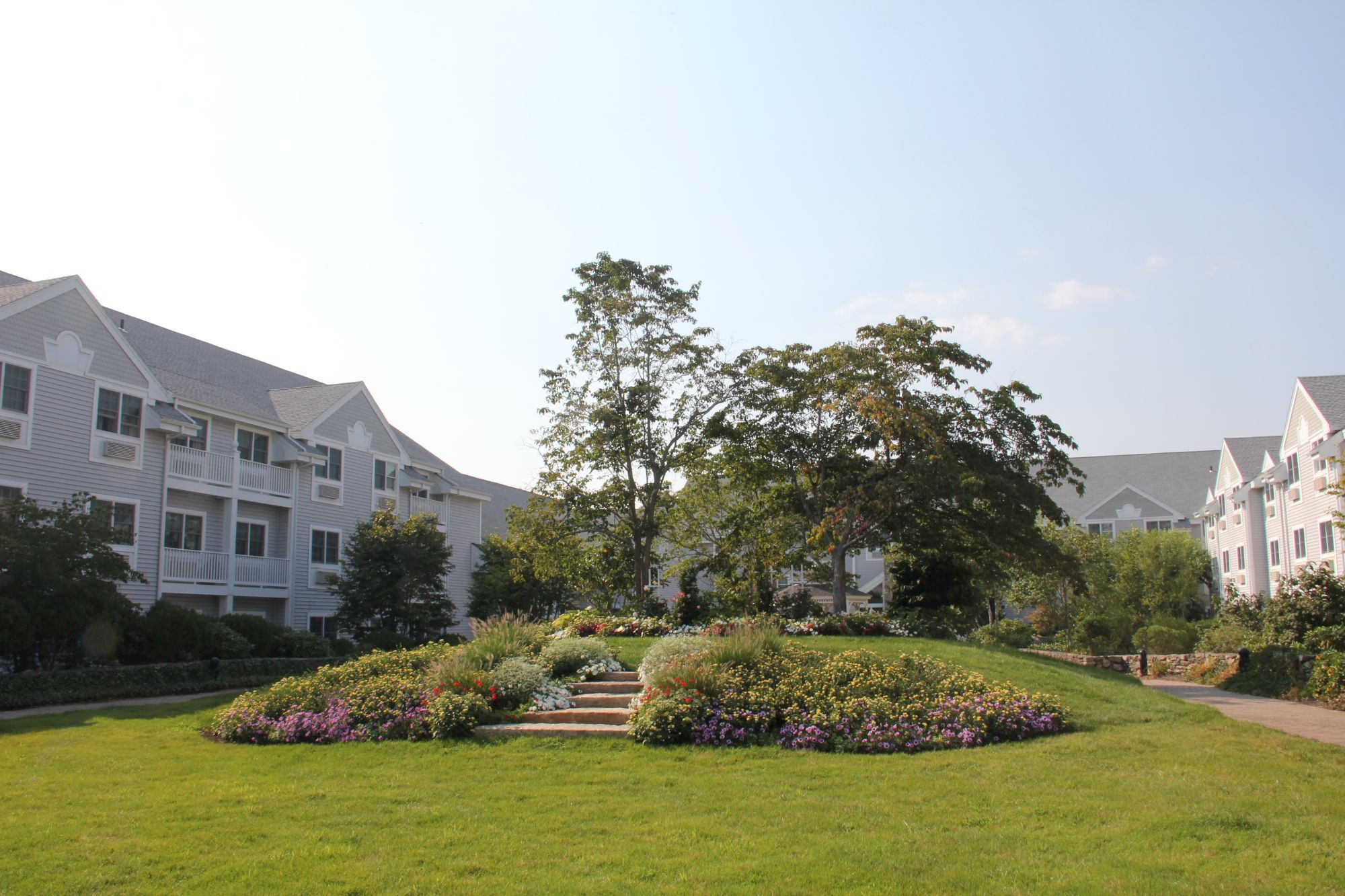 A landscaped courtyard with circular flower beds, green lawn, and trees surrounded by white multi-story apartment buildings, sunny day.