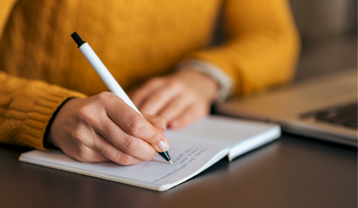 A person in a mustard sweater writes in a notebook with a pen beside a laptop, focused on jotting notes.