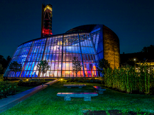 A modern glass-building theater lit in blue at night, with a curved glass facade, surrounding garden, and a glowing tower in the background.