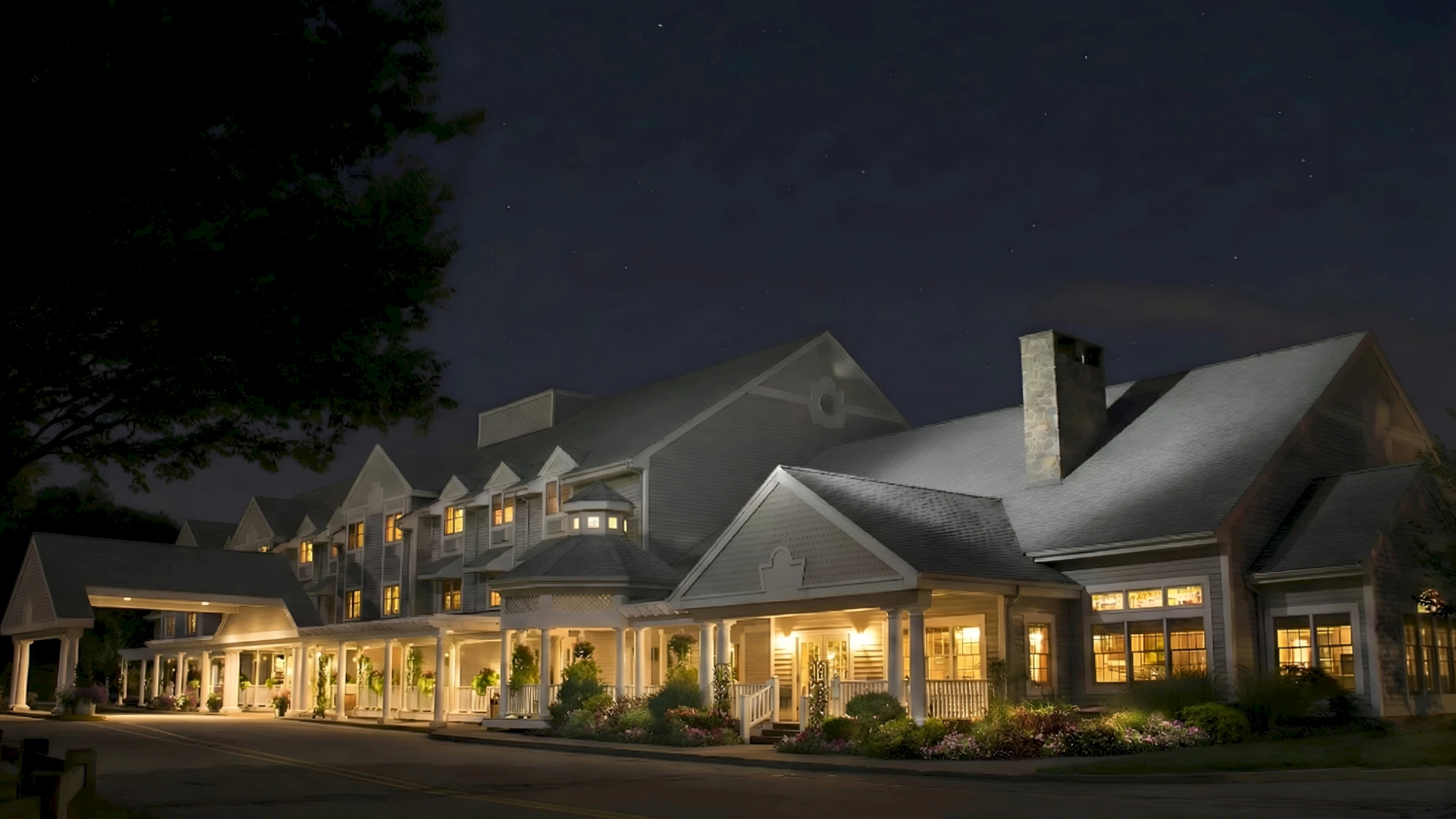 A large, elegant hotel at night with warmly lit windows and a welcoming front porch, surrounded by landscaping and a dark sky.