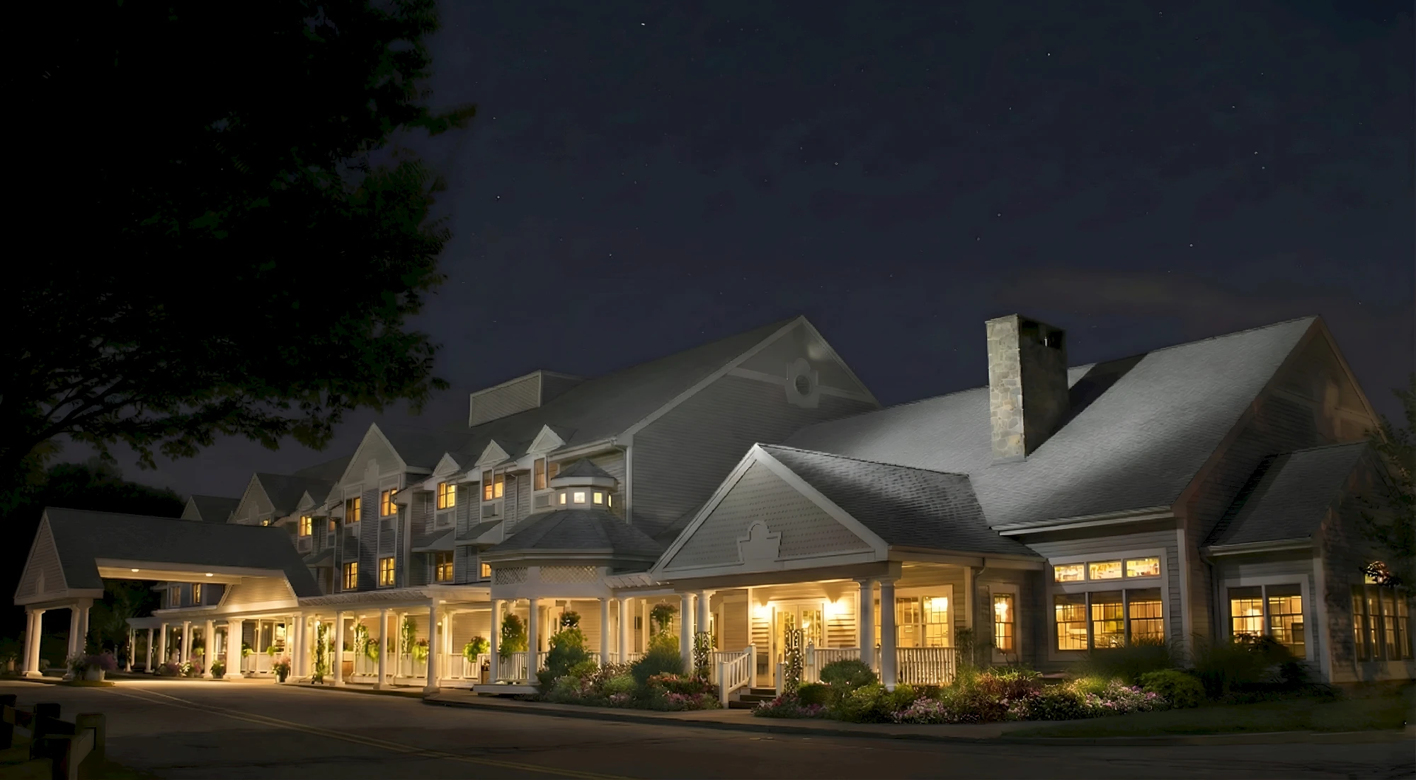 A large, elegant hotel at night with warmly lit windows and a welcoming front porch, surrounded by landscaping and a dark sky.