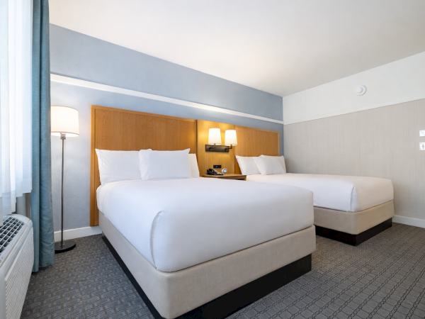 Two double beds with white linens in a modern hotel room, wooden headboard, a nightstand lamp between beds, a floor lamp by the window, and a blue-gray color scheme.