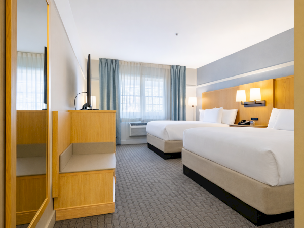 A hotel room with two queen beds, a desk and mirror, a window with blue curtains, a nightstand lamp, and neutral gray carpet.