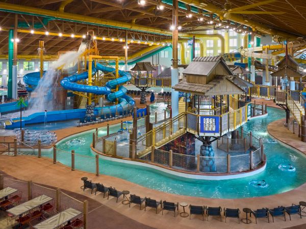 A large indoor water park with colorful slides, a water turret, lazy river, and seating areas under a high, lit wooden roof.