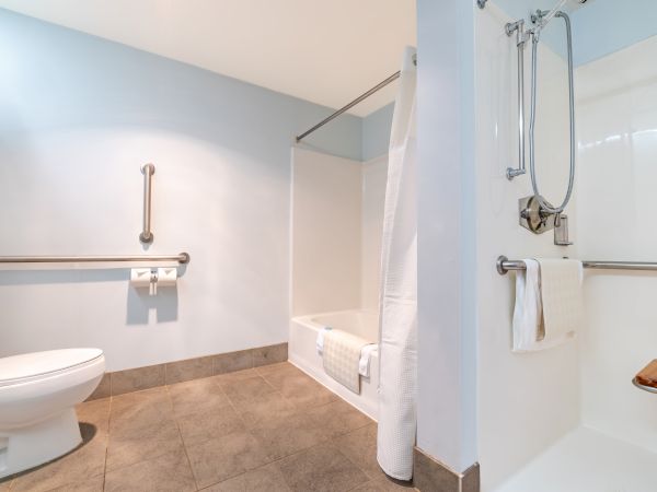 A modern bathroom with a toilet, a walk-in shower with a shower curtain, a bathtub, grab bars for accessibility, and light blue walls.