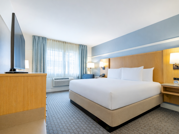 A modern hotel room with a large bed, wooden headboard, wall lamps, a dresser with a TV, a window with blue curtains, and a small seating area.
