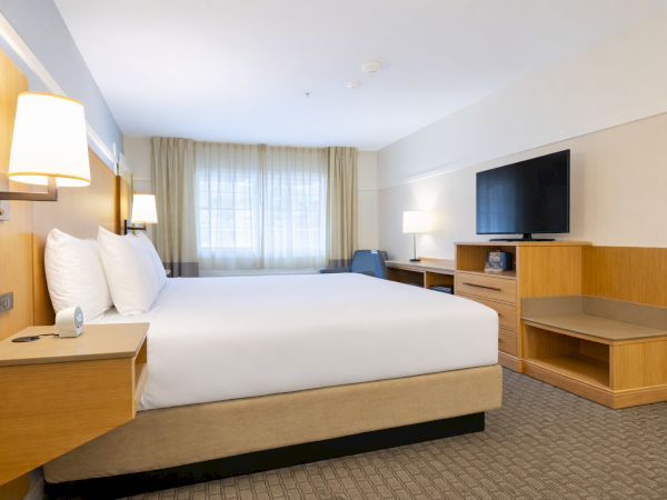 A modern hotel room with a large bed, bedside lamps, a desk, a wall-mounted TV, a chair near a window, and neutral-toned decor for a calm, functional space.