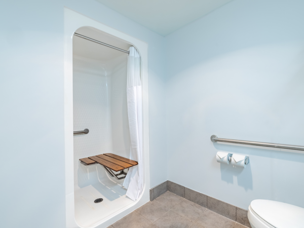 A light blue-accessible bathroom with a built-in shower stall, wooden shower seat, grab bars, toilet, and toilet paper holder along the wall.