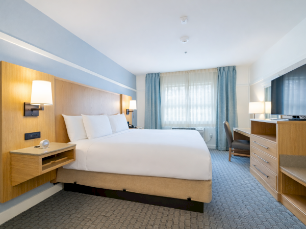 A modern hotel room with a large bed, wood headboard, bedside lamps, a desk, and a flat-screen TV; soft blue accents and a window with curtains.