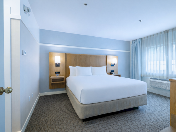 A tidy hotel room with a king bed, blue walls, built-in headboard, nightstands, and a large window with sheer curtains, plus a dresser to the right.