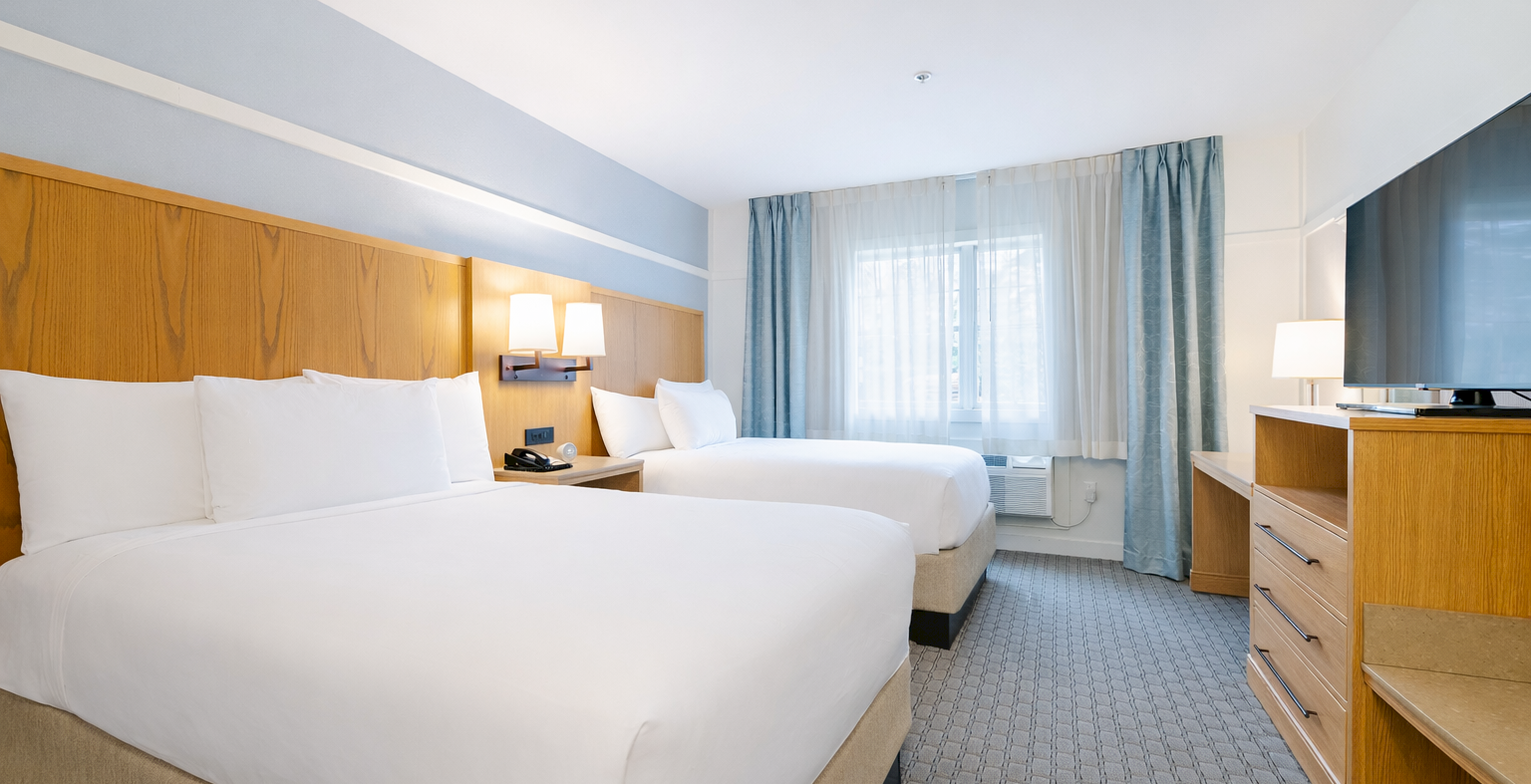 Two clean hotel beds with white linens, wood headboard, bedside lamps, a desk, dresser, and a window with blue curtains in a bright room.