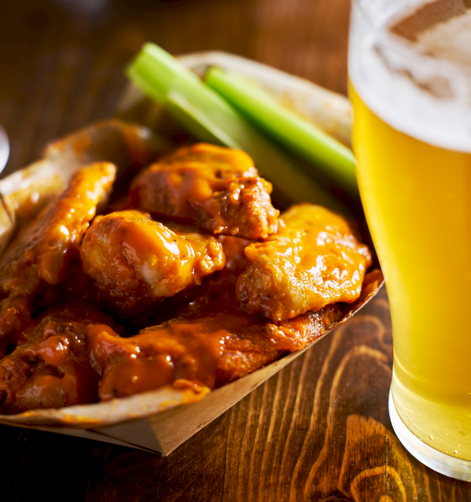 Crispy buffalo wings with dipping sauce and celery sticks beside a frosty beer in a rustic setting.