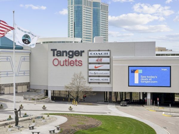 A view of Tanger Outlets with store signs and flags, plus the &ldquo;Tanger&rdquo; tower rising under a blue sky.